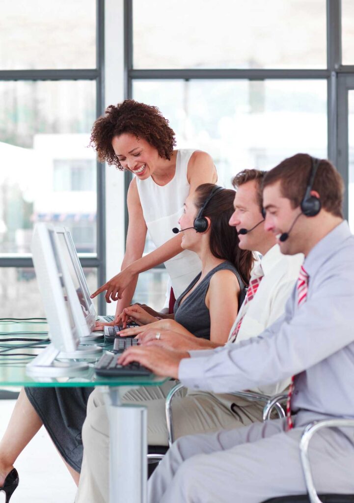 A cheerful answering service team member assisting colleagues with customer inquiries on computers in a bright office environment, embodying teamwork and support